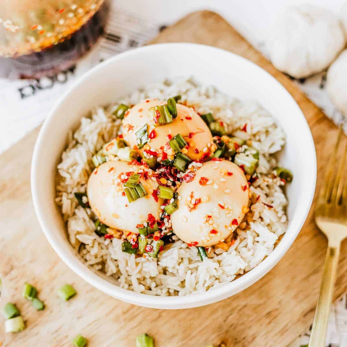 A white bowl filled with rice topped with three Korean marinated eggs, garnished with chopped green onions, sesame seeds, and red chili flakes, placed on a wooden board with a fork nearby.