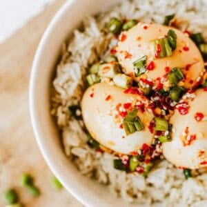 A bowl of white rice topped with three soy sauce marinated eggs, garnished with chopped green onions, red chili flakes, and sesame seeds.