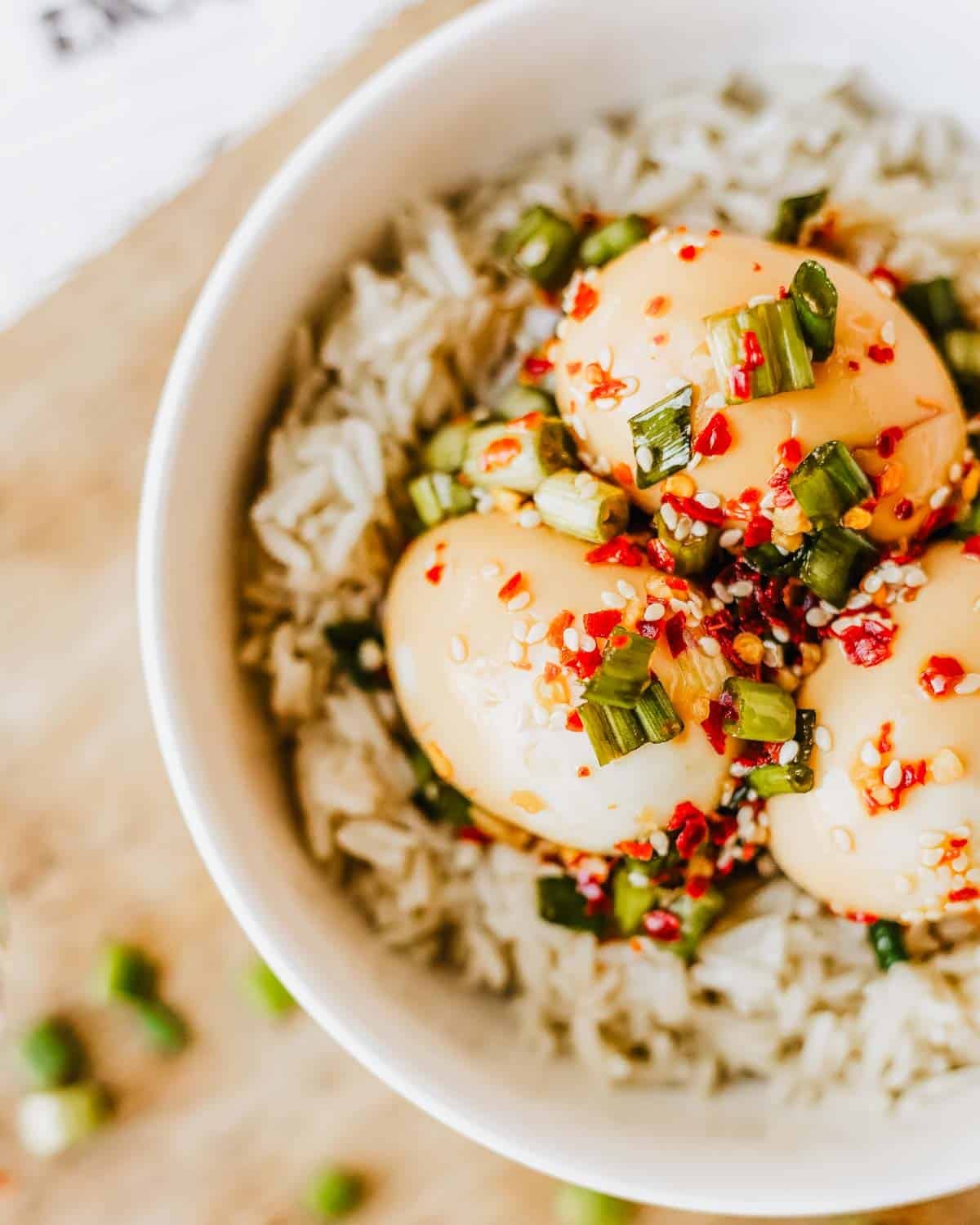 A bowl of white rice topped with three soy sauce marinated eggs, garnished with chopped green onions, red chili flakes, and sesame seeds.