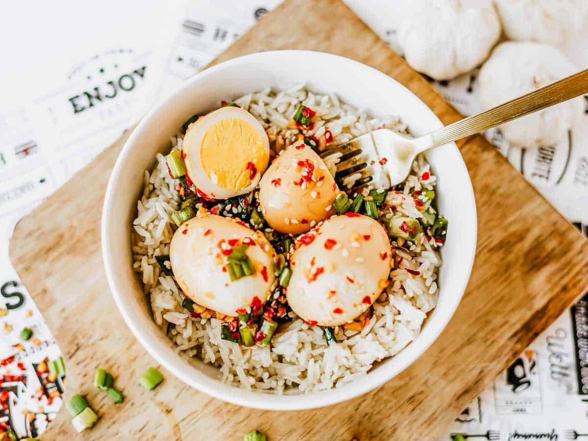 A bowl of rice topped with three Korean marinated eggs, sliced green onions, and red chili flakes, with a fork resting on the side, placed on a wooden board.