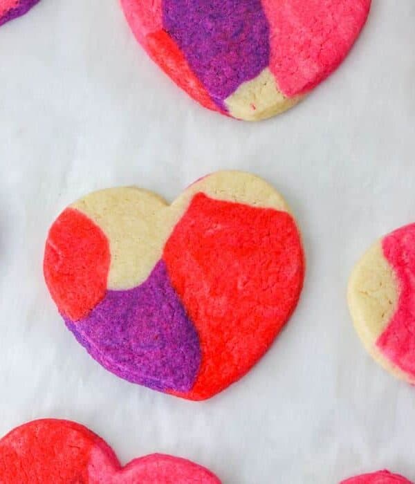 Heart-shaped classic sugar cookies with a marbled pattern in pink, red, purple, and plain dough are arranged on a white surface.