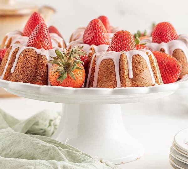A round bundt cake topped with whole strawberries and drizzled with white icing sits on a white cake stand. Plates, a teapot, and a green cloth are nearby on a bright, elegant table setting.