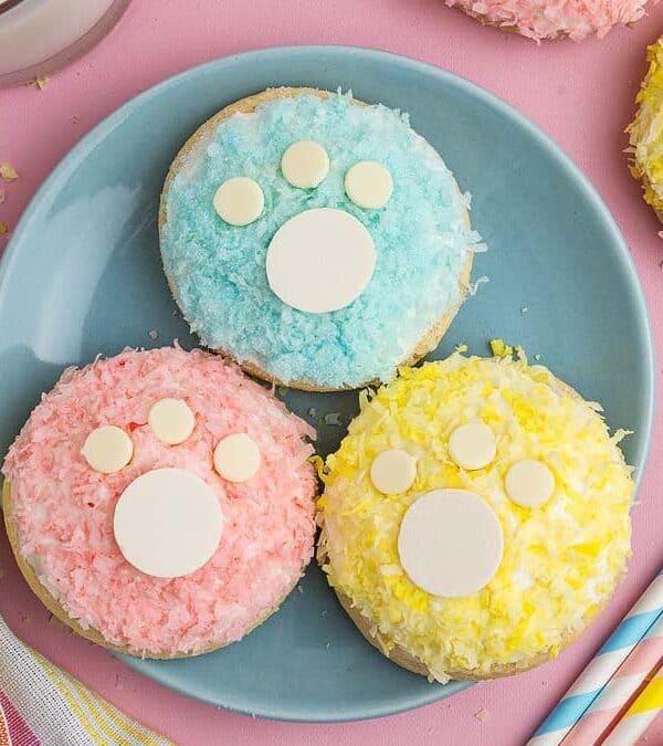 Three adorable cookies in pastel pink, blue, and yellow with paw print designs sit on a blue plate. Two striped straws and a colorful dish towel are nearby on a pink surface, with a glass of milk partially visible.