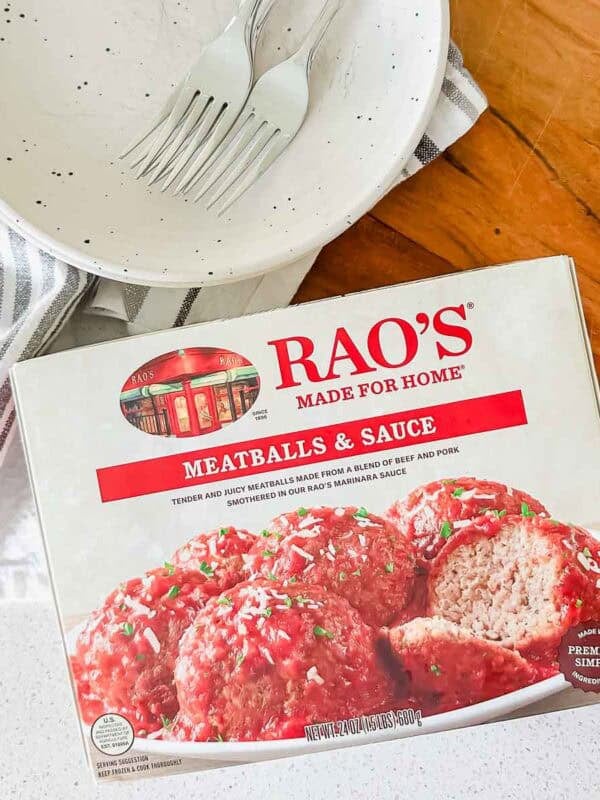 A box of Rao's Meatballs & Sauce sits on a counter next to an empty plate with two forks, a striped napkin, and part of a wooden table visible in the background.