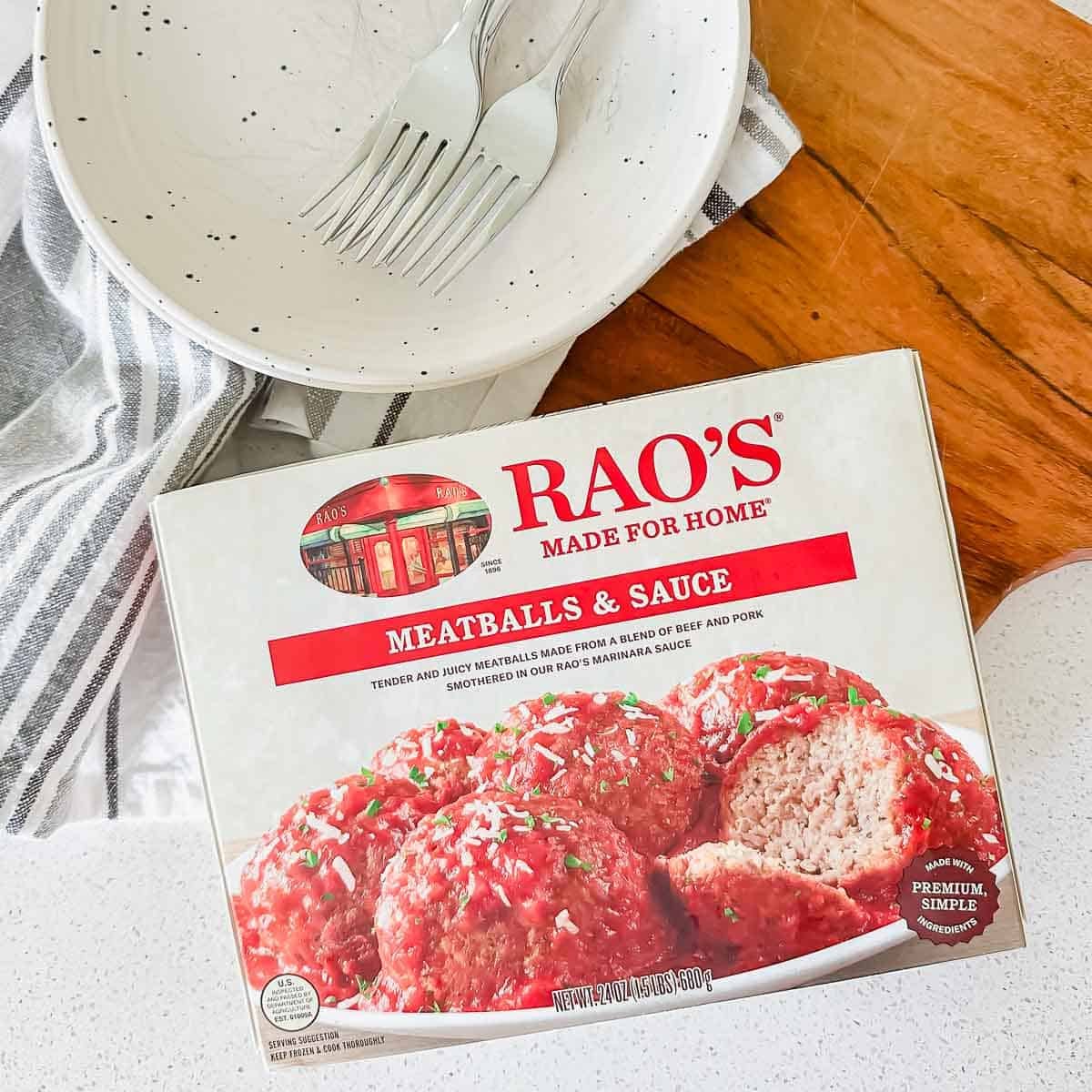 A box of Rao's Meatballs & Sauce sits on a counter next to an empty plate with two forks, a striped napkin, and part of a wooden table visible in the background.