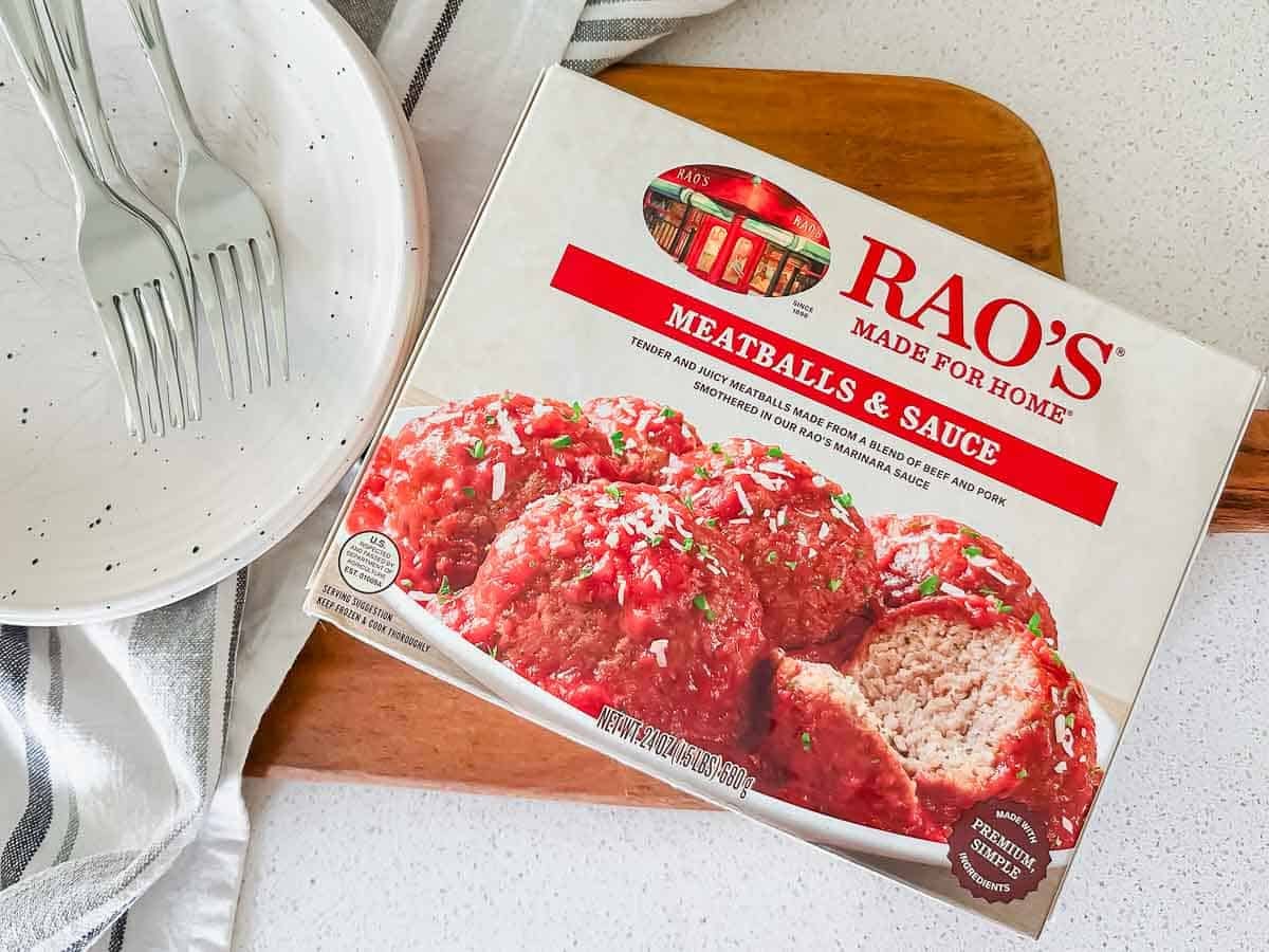 A box of Rao's Meatballs & Sauce sits on a wooden cutting board next to a white plate with black speckles and three forks, with a striped towel underneath. The box shows an image of meatballs covered in tomato sauce.