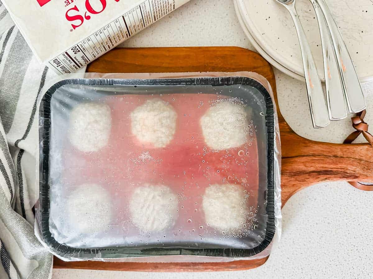 A frozen entree of six meatballs in sauce covered with plastic wrap sits on a wooden board next to a striped towel, a carton, a plate, and two metal forks.