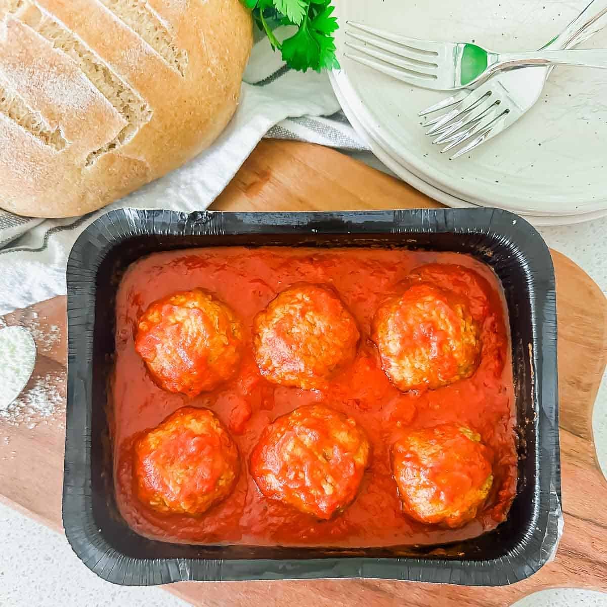 A black frozen dinner tray with six meatballs in tomato sauce sits on a wooden board, next to a loaf of bread, stacked white plates, and forks. Fresh herbs are partly visible in the background.