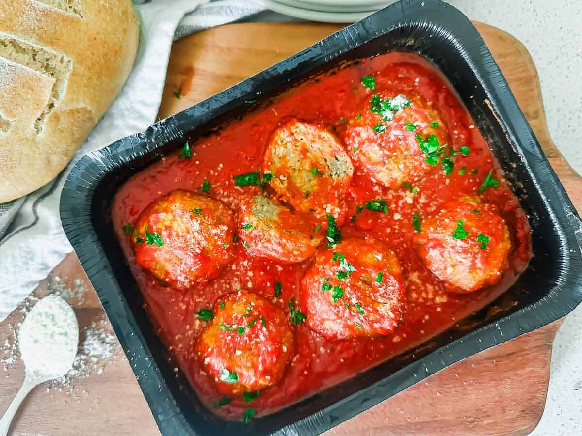 A frozen dinner tray filled with six meatballs in marinara sauce, garnished with chopped parsley and grated cheese. The tray is on a wooden board next to a loaf of bread and a spoon with seasoning.
