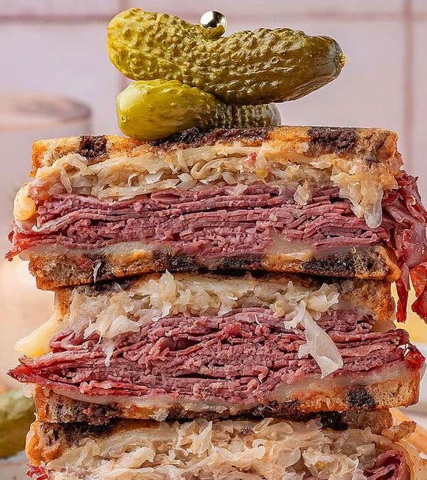 A stacked Reuben sandwich filled with layers of corned beef, sauerkraut, and melted cheese between toasted rye, topped with three small pickles on a skewer. One of the best sandwiches for lunch recipes—served with chips and a drink in the background.