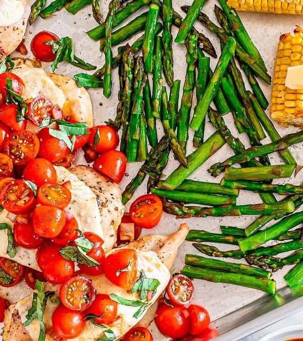Sheet pan with baked chicken breasts topped with melted cheese, sliced cherry tomatoes, and basil, alongside roasted asparagus spears and corn on the cob with pats of butter.