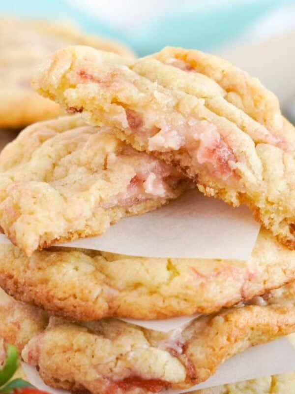A stack of strawberry cookies separated by parchment paper, with the top cookie broken in half to reveal a soft, gooey center—perfect for strawberry desserts or sweet strawberry recipes. Fresh strawberries and more cookies are visible in the background.