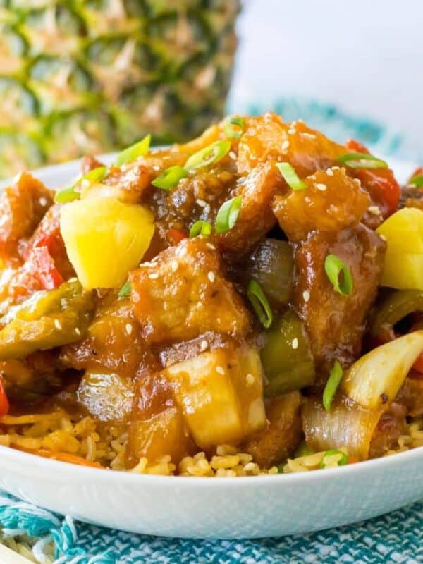 A bowl of sweet and sour stir-fry with vegetables, pineapple chunks, and sauce served over rice, garnished with green onions—a perfect effortless dinner inspired by pork slow cooker recipes, with a whole pineapple in the background.