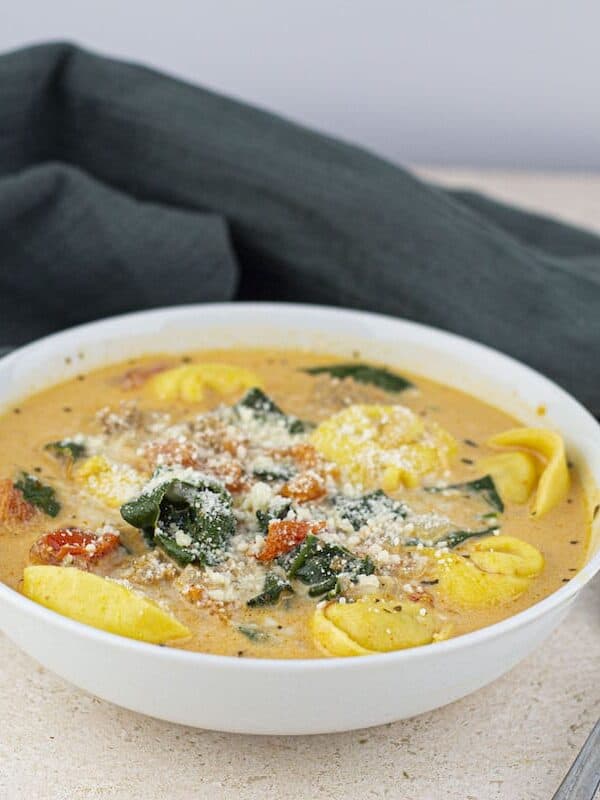A bowl of creamy tortellini soup with spinach and diced tomatoes, sprinkled with grated cheese, showcases a comforting classic from the world of Slow Cooker Soups, sitting beside a spoon and a dark green napkin.