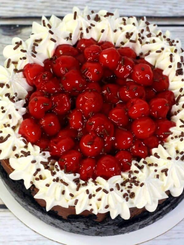 A round chocolate cake topped with whipped cream, chocolate sprinkles, and a generous layer of glazed red cherries, displayed on a white plate over a wooden surface.