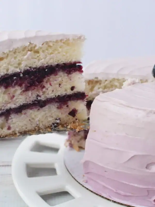 A slice of layered cake with light purple frosting and a blueberry filling is being lifted from the whole celebration cake, which is topped with fresh blueberries and sits on a white plate.