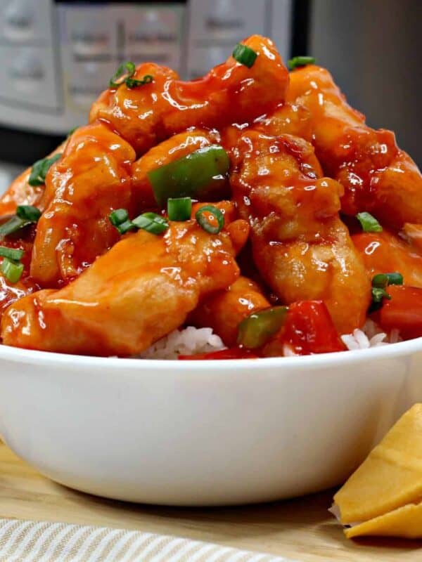 A white bowl filled with orange-glazed chicken and green peppers over white rice, surrounded by four fortune cookies. An Instant Pot and stacked plates in the background hint at easy Instant Pot chicken recipes for quick weeknight dinners.