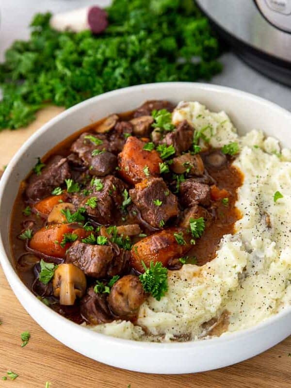 A bowl of beef stew with carrots and mushrooms in a rich sauce, served alongside creamy mashed potatoes, garnished with chopped parsley. Forks, fresh herbs, and an Instant Pot are nearby—perfect comfort food for cozy weeknight dinners.