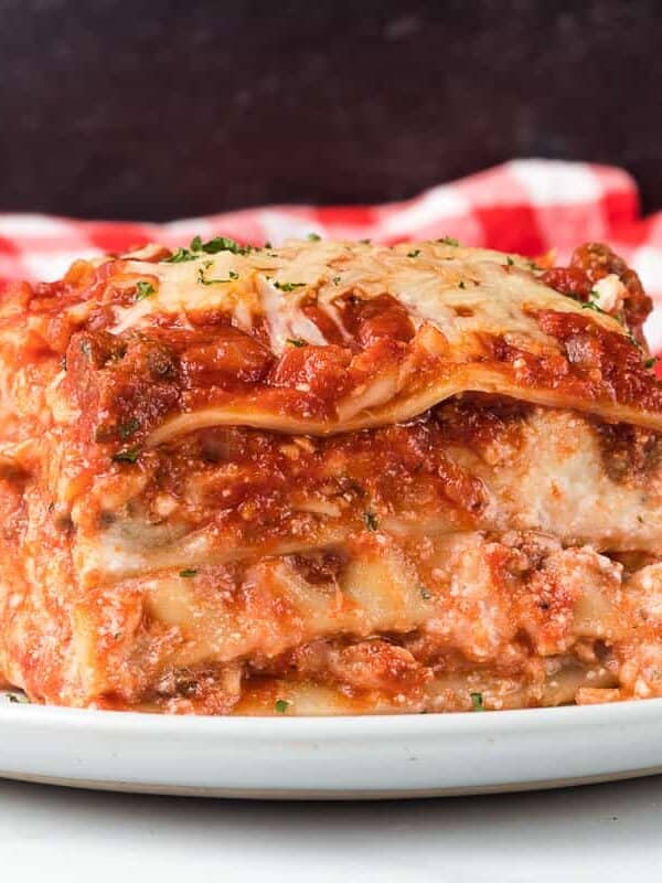 A slice of lasagna with layers of pasta, cheese, and tomato sauce sits on a white plate, garnished with herbs, in front of a red and white checkered cloth.
