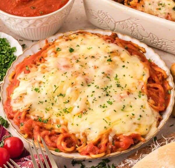 A plate of baked spaghetti topped with melted cheese and herbs, surrounded by breadsticks, grated cheese, chopped parsley, cherry tomatoes, and bowls of sauce on a light-colored table.