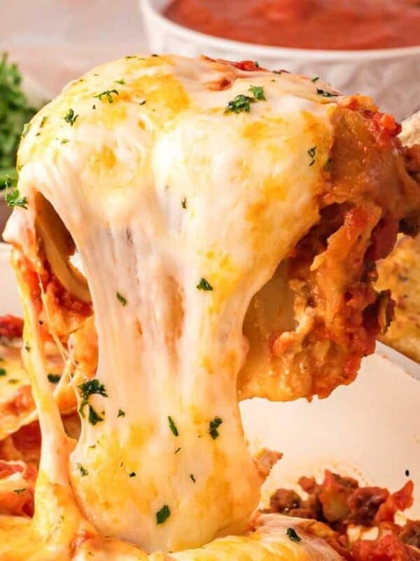 A serving of easy lasagna being lifted from a dish, with melted cheese stretching, surrounded by salad, garlic bread, and a bowl of tomato sauce—perfect for trying new lasagna recipes.