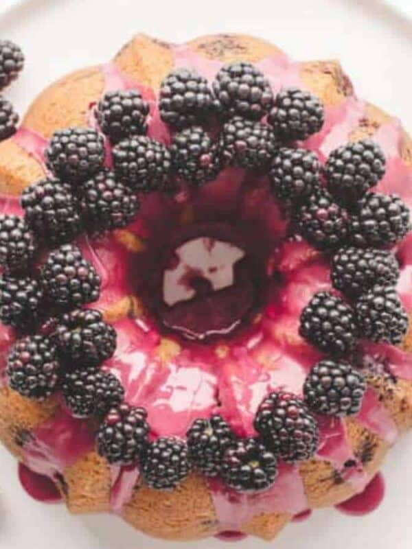 A Bundt cake inspired by bright lemon cake recipes, topped with glossy purple icing and fresh blackberries, is beautifully displayed on a white plate.