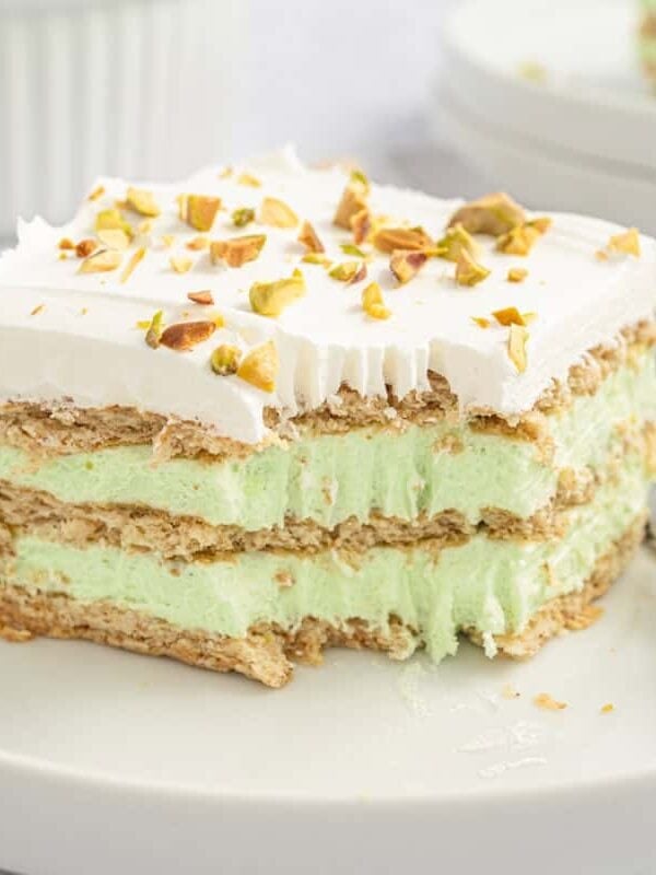 A No-Bake Icebox Cake with layers of graham crackers, light green pistachio cream, and whipped topping is served on a white plate, garnished with chopped pistachios. A fork with a bite-sized piece rests beside this perfect summer dessert.