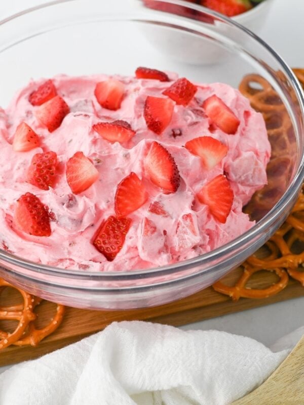 A glass bowl brimming with pink strawberry fluff salad, one of the best easy dessert recipes, is topped with fresh strawberries and sits on a wooden board lined with pretzels. A wooden spoon and white cloth add charm, with more strawberries in the background.