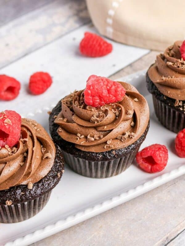 Three chocolate cupcakes topped with swirls of chocolate frosting and a fresh raspberry, displayed on a white rectangular plate with scattered raspberries—an irresistible treat for lovers of rich desserts and decadent chocolate recipes.