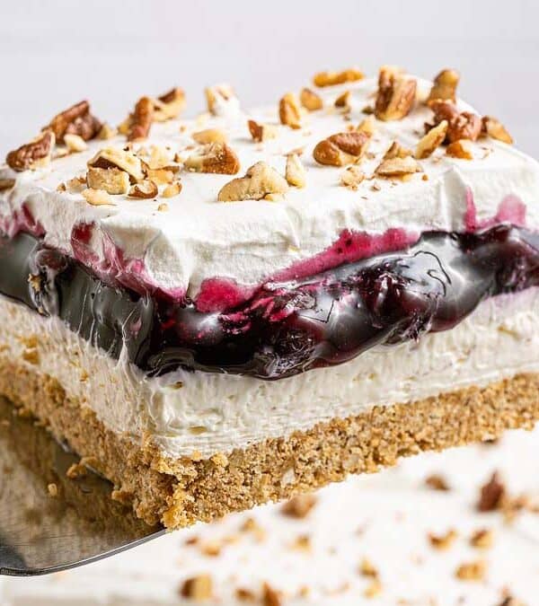 A square slice of lush dessert with a graham cracker crust, creamy filling, blueberry topping, and whipped cream, garnished with chopped pecans, held on a spatula above a dish. Bowls of nuts and blueberries are in the background.