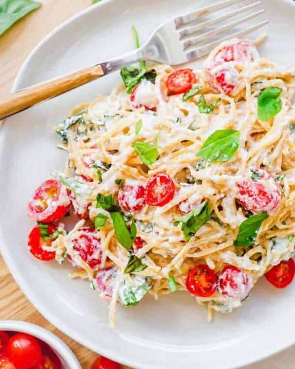 A plate of creamy pasta topped with halved cherry tomatoes and fresh basil leaves, with a fork resting on the plate. Extra basil and cherry tomatoes are scattered around—perfect for those seeking delicious takeout alternatives or new dinner ideas.