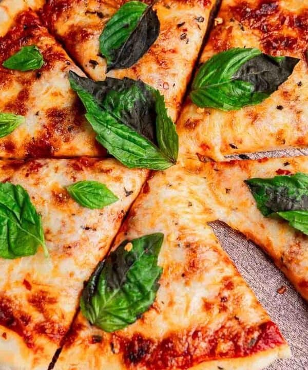 A cheese pizza cut into eight slices, topped with fresh basil leaves, sits on a rustic wooden surface—perfect for those who love crispy recipes made with simple ingredients.