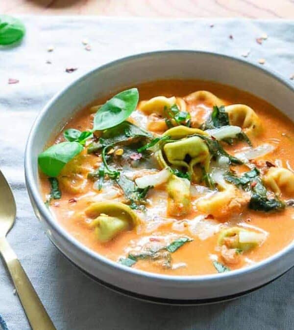 A bowl of creamy tortellini soup with spinach and herbs sits on a cloth napkin, next to a gold spoon. This comfort food features an orange broth, green tortellini, and garnish. A box of vegan cheese is partially visible nearby.
