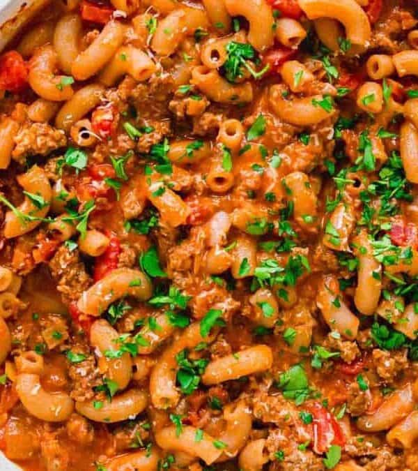 A skillet filled with cooked macaroni pasta, ground beef, diced tomatoes, and a creamy sauce, garnished with chopped fresh parsley. A wooden spoon rests nearby on a striped kitchen towel.