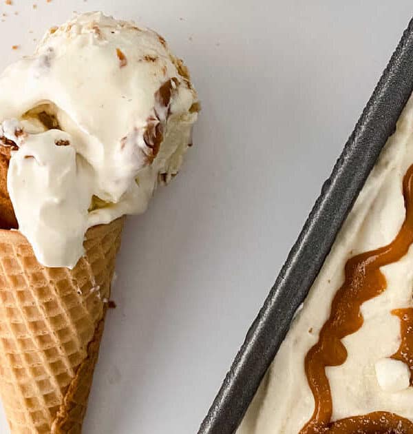 A waffle cone with a scoop of vanilla ice cream and a cookie piece is next to a tray of ice cream topped with caramel sauce and cookies. Crumbled cookies are scattered on a white surface.