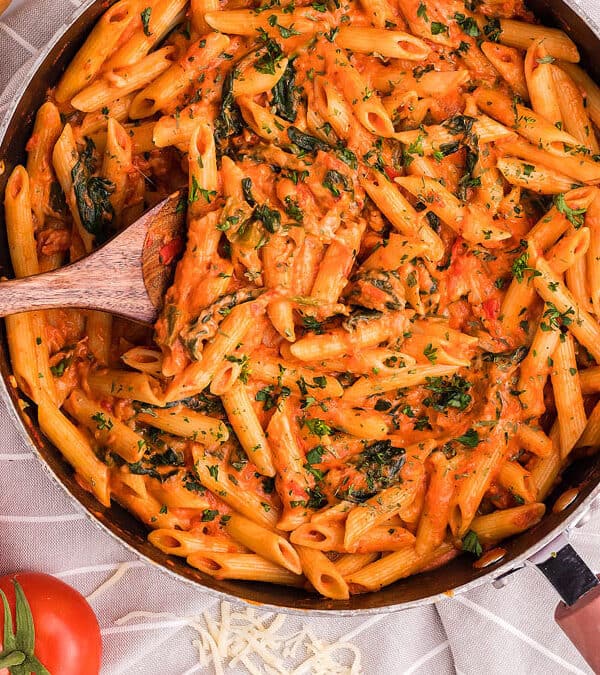A skillet filled with creamy tomato penne pasta, a favorite in Italian cuisine, garnished with chopped parsley. Around it are fresh tomatoes, spinach leaves, shredded cheese, a green bell pepper, and a baguette. A wooden spoon rests in the pasta.