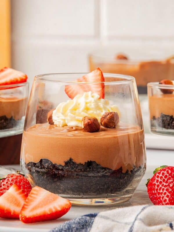Three dessert glasses filled with layered chocolate mousse, topped with whipped cream, sliced strawberries, and hazelnuts. Perfect for fans of Nutella desserts, with fresh strawberries and nuts scattered around on a white surface.