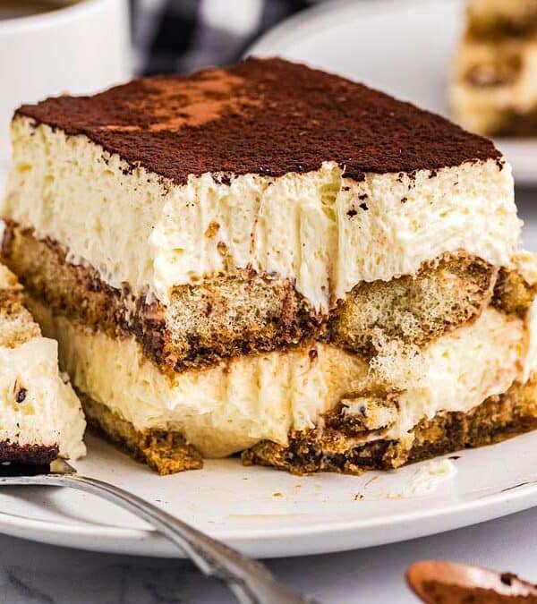 A slice of tiramisu with creamy layers and cocoa powder on top sits on a white plate, with a forkful removed. A cup of coffee and more dessert are blurred in the background.
