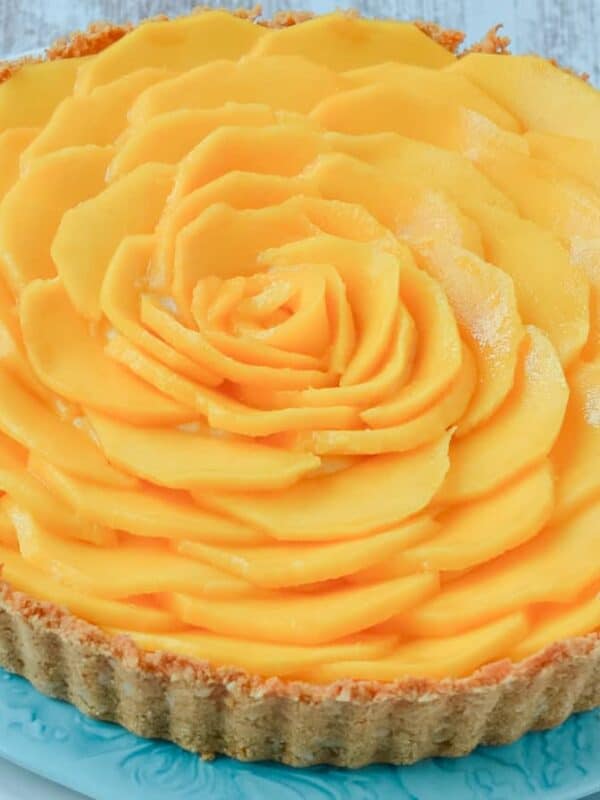 A tart with a crumbly crust filled and beautifully topped with thinly sliced mango arranged in overlapping circles to resemble a flower, placed on a blue plate.