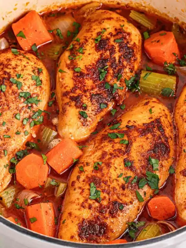 A white pot filled with four seasoned chicken breasts, cooked with chopped carrots and celery in a tomato-based sauce, garnished with fresh herbs. Perfect for comforting recipes and a delicious takeout alternative. Parsley and a checkered napkin are beside the pot.