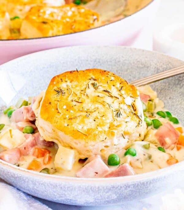 A bowl of creamy chicken pot pie filling with leftover ham, peas, and carrots, topped with a golden-brown biscuit. A spoon rests in the bowl, and a pink baking dish is visible in the background—perfect for creative recipes using ham.