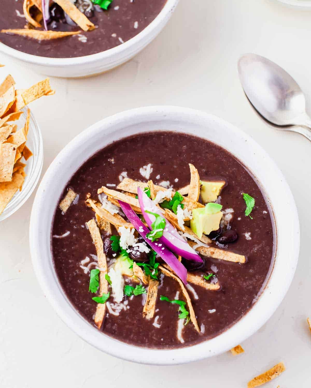 A bowl of Instant Pot Black Bean Soup topped with tortilla strips, avocado, red onion, grated cheese, cilantro, and black beans, served with a spoon and extra tortilla strips on the side.