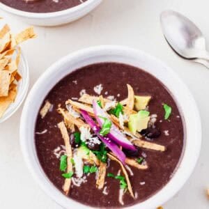 A bowl of Instant Pot Mexican black bean soup topped with tortilla strips, avocado, red onion, black beans, cheese, and chopped cilantro, with a spoon and extra tortilla strips on the side.