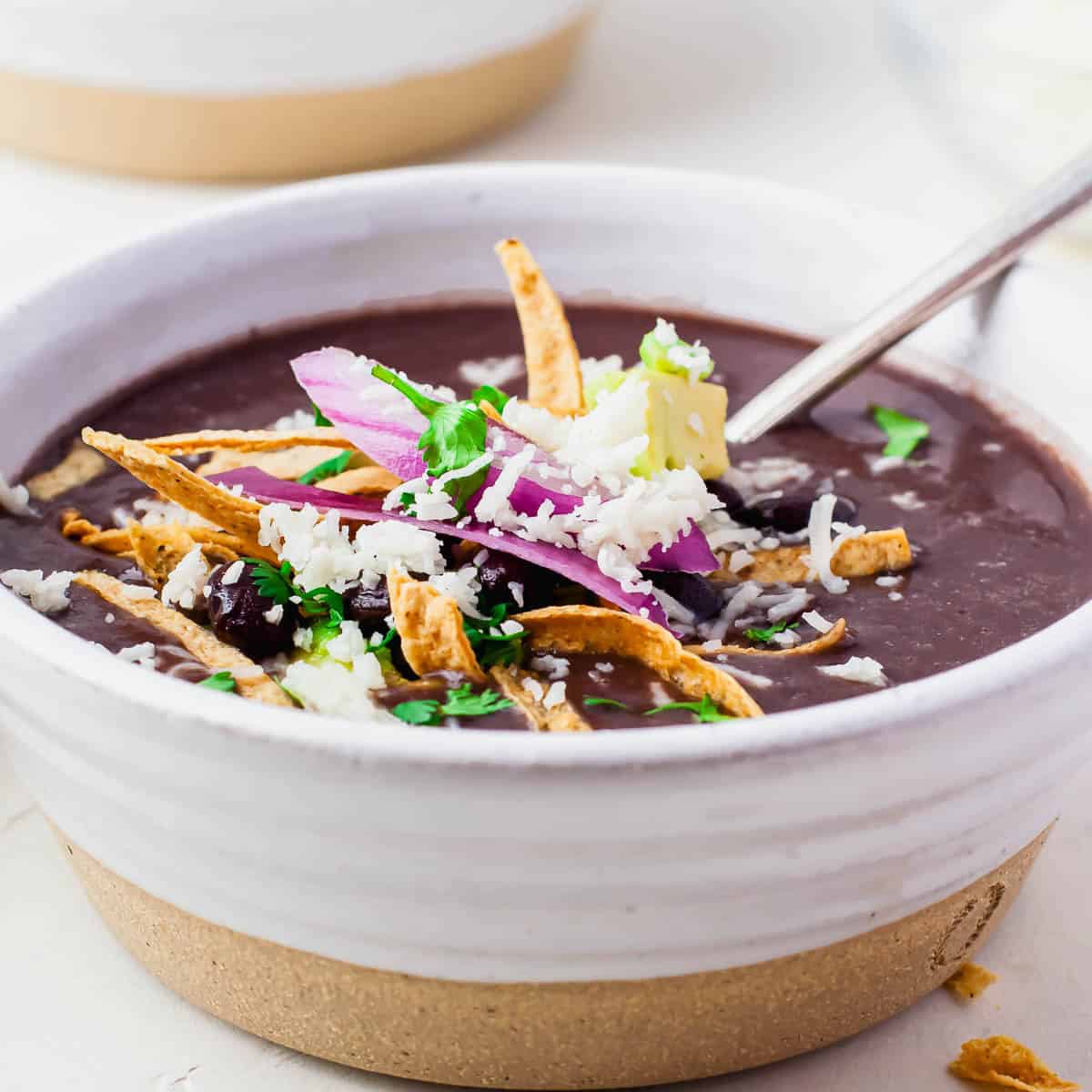 A bowl of black bean soup topped with shredded cheese, crispy tortilla strips, sliced red onion, avocado, and cilantro, with a spoon resting inside the bowl.
