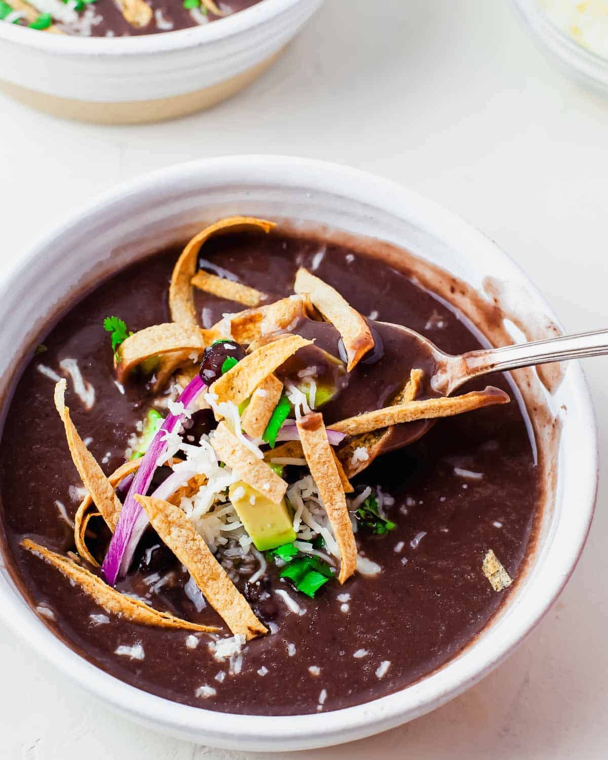 A bowl of black bean soup made in the Instant Pot topped with crispy tortilla strips, shredded cheese, red onion, cilantro, and avocado, with a spoon lifting a portion from the bowl.
