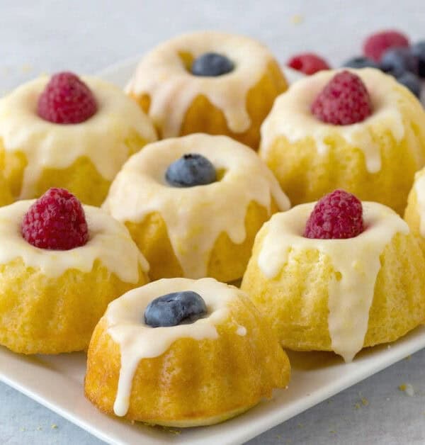 A plate of mini bundt cakes topped with white icing, each garnished with a fresh blueberry or raspberry. A few loose berries are scattered in the background.