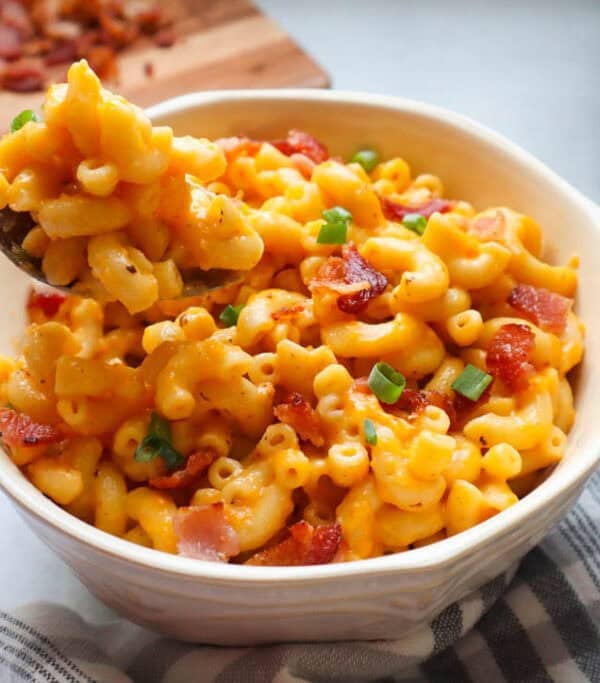 A bowl of creamy macaroni and cheese topped with crispy bacon bits and chopped green onions, with a spoon lifting a portion—an irresistible choice for weeknight dinners. A cutting board with bacon and a kitchen appliance are in the background.