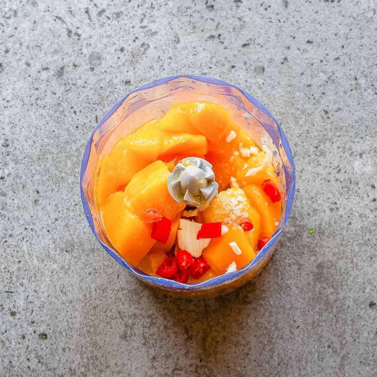 A food processor bowl containing chunks of orange mango, red chili slices, and pieces of garlic, sitting on a gray textured surface.