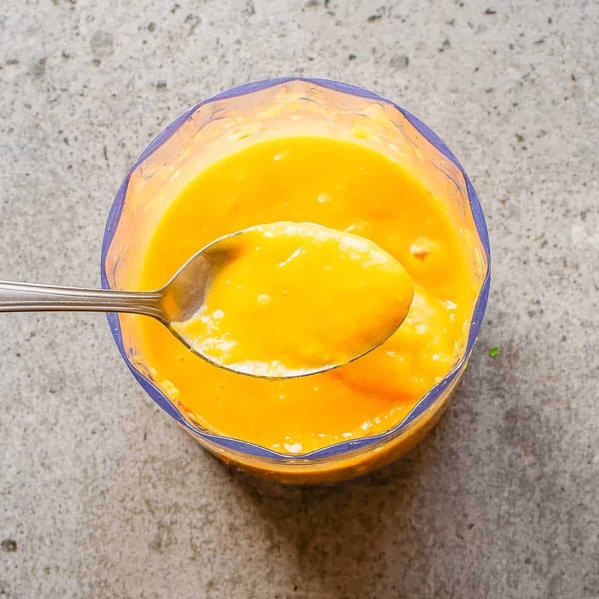 A spoon holding thick, bright yellow mango sauce over a clear glass filled with more of the puree, set on a light gray textured surface.