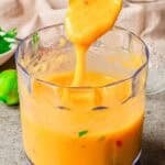 A spoon lifts thick, creamy mango sauce from a clear food processor bowl. Some green and red flecks are visible in the sauce. The background shows a lime, herbs, and a glass jar on a gray countertop.